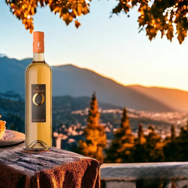 Photo d'une bouteille de vin blanc 'Zeste d'O' des Vignobles Jourdain posée sur une table en bois avec une nappe rouge. En arrière-plan, un paysage de montagnes et d'arbres au coucher du soleil, créant une ambiance chaleureuse et pittoresque